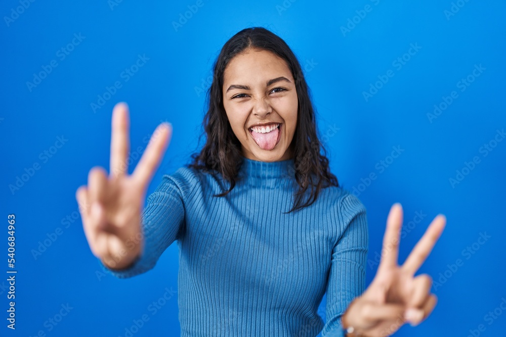 Young brazilian woman standing over blue isolated background smiling with tongue out showing fingers of both hands doing victory sign. number two. Young brazilian woman standing over blue isolated background smiling with tongue out showing fingers of both hands doing victory sign. number two.