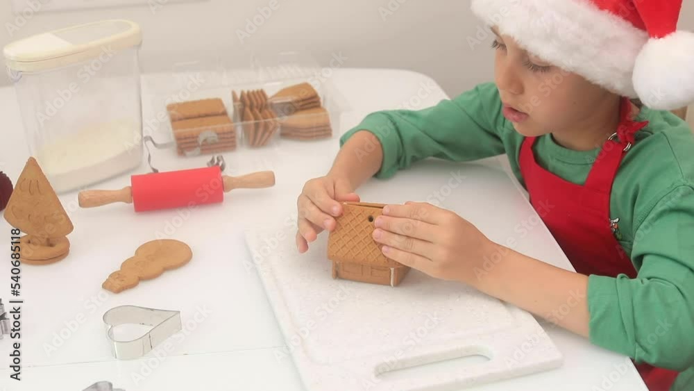 A child in a Claus hat is making a gingerbread house in the kitchen. A child in a Claus hat is making a gingerbread house in the kitchen.