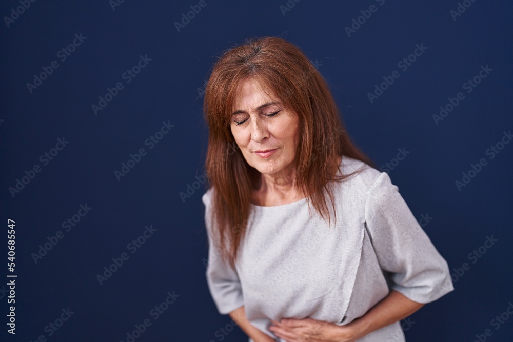 Middle age woman standing over blue background with hand on stomach because indigestion, painful illness feeling unwell. ache concept.