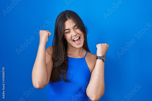 Hispanic woman standing over blue background celebrating surprised and amazed for success with arms raised and eyes closed. winner concept.