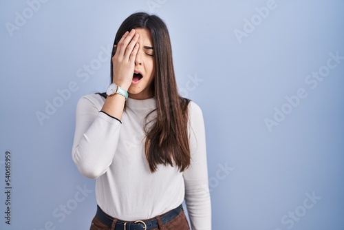 Young brunette woman standing over blue background yawning tired covering half face, eye and mouth with hand. face hurts in pain.