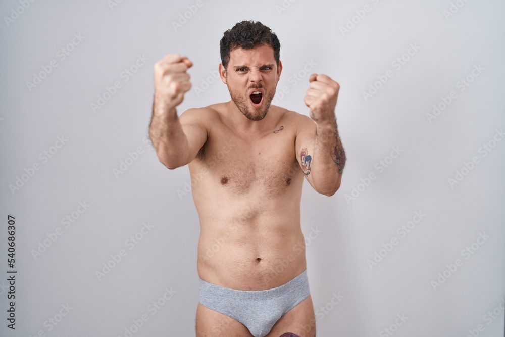 Young hispanic man standing shirtless wearing underware angry and mad raising fists frustrated and furious while shouting with anger. rage and aggressive concept.