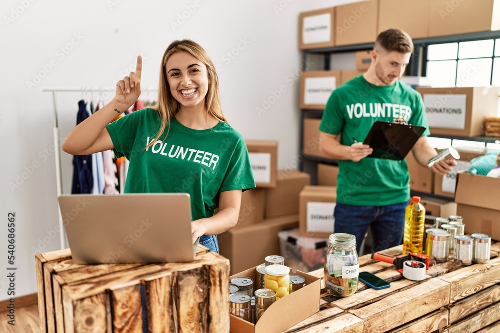 Young woman and man wearing volunteer t shirt at donations stand surprised with an idea or question pointing finger with happy face, number one Young woman and man wearing volunteer t shirt at donations stand surprised with an idea or question pointing finger with happy face, number one