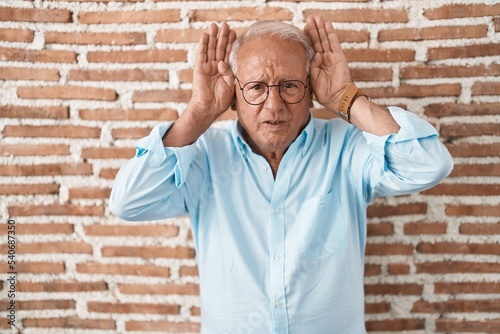 Senior man with grey hair standing over bricks wall doing bunny ears gesture with hands palms looking cynical and skeptical. easter rabbit concept. photo