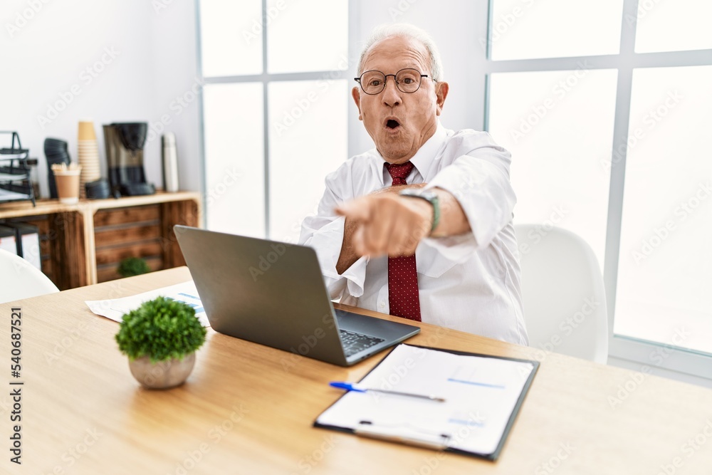Senior man working at the office using computer laptop pointing with finger surprised ahead, open mouth amazed expression, something on the front Senior man working at the office using computer laptop pointing with finger surprised ahead, open mouth amazed expression, something on the front