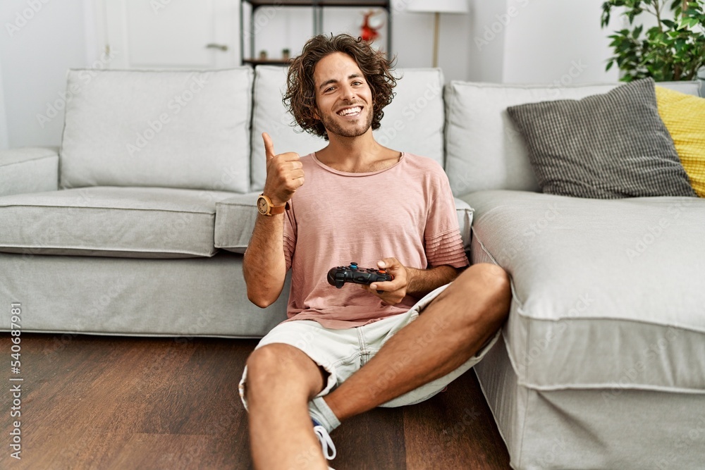 Young hispanic man playing video game holding controller at home smiling happy and positive, thumb up doing excellent and approval sign