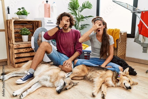 Photography Young hispanic couple doing laundry with dogs peeking in shock covering face and eyes with hand, looking through fingers with embarrassed expression