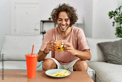Young hispanic man smiling happy eating classic burger and drinking soda at home.