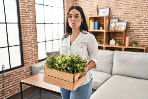 Young hispanic woman holding green plants at the living room making fish face with mouth and squinting eyes  crazy and comical.