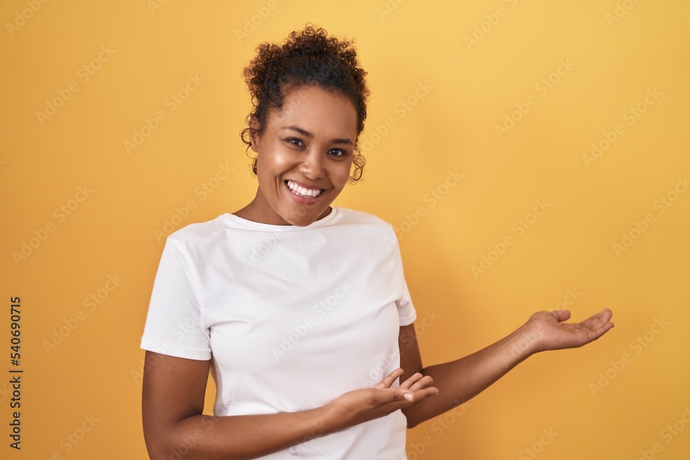 Young hispanic woman with curly hair standing over yellow background inviting to enter smiling natural with open hand Young hispanic woman with curly hair standing over yellow background inviting to enter smiling natural with open hand