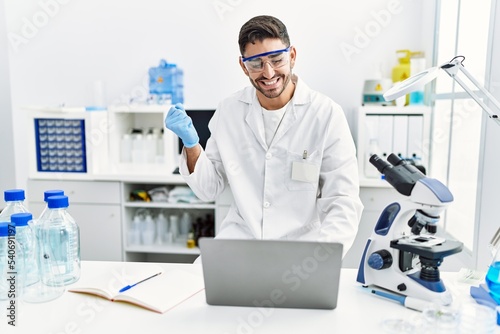 Young hispanic man working at scientist laboratory with laptop screaming proud  celebrating victory and success very excited with raised arm
