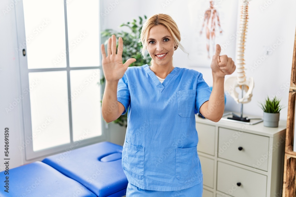 Beautiful blonde physiotherapist woman working at pain recovery clinic showing and pointing up with fingers number six while smiling confident and happy. Beautiful blonde physiotherapist woman working at pain recovery clinic showing and pointing up with fingers number six while smiling confident and happy.