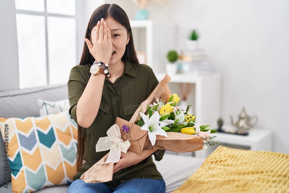 Chinese young woman holding bouquet of white flowers covering one eye with hand, confident smile on face and surprise emotion.