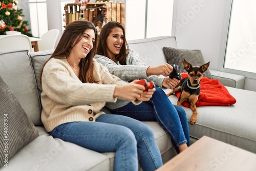 Two women playing video game sitting with dog by christmas tree at home
