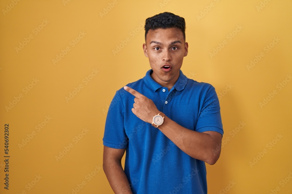 Young hispanic man standing over yellow background surprised pointing with finger to the side, open mouth amazed expression. Young hispanic man standing over yellow background surprised pointing with finger to the side, open mouth amazed expression.