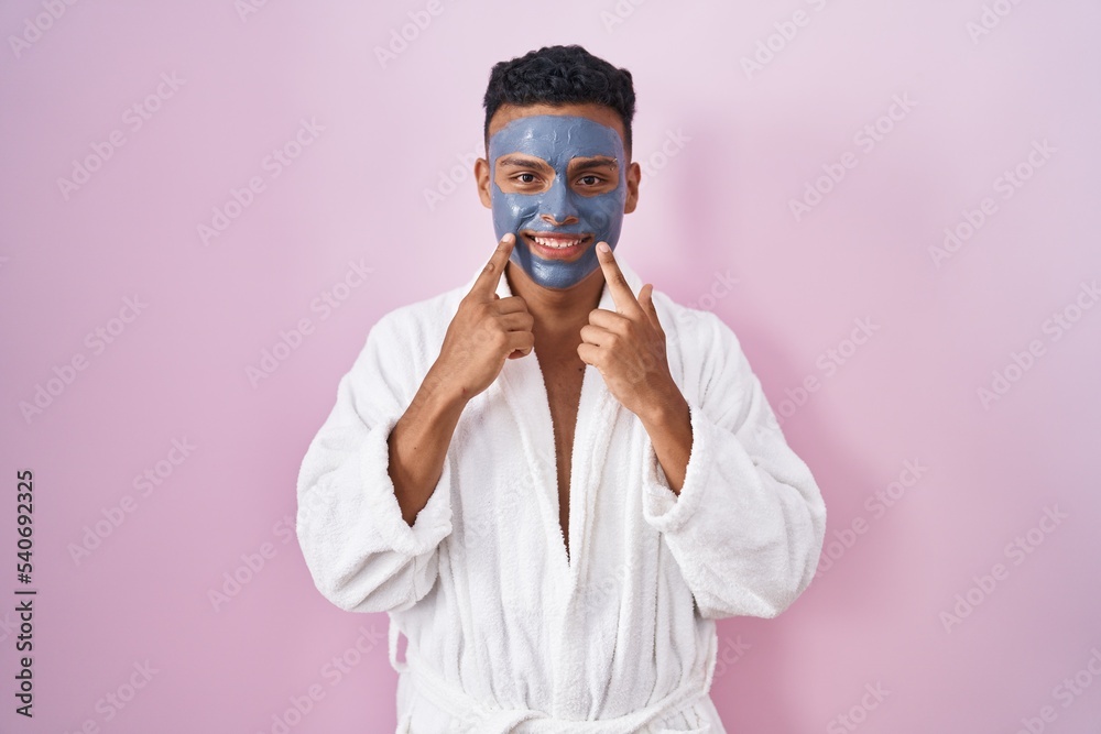 Young hispanic man wearing beauty face mask and bath robe smiling with open mouth, fingers pointing and forcing cheerful smile Young hispanic man wearing beauty face mask and bath robe smiling with open mouth, fingers pointing and forcing cheerful smile