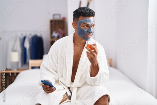 Young latin man sitting on bed using smartphone drinking whisky at bedroom