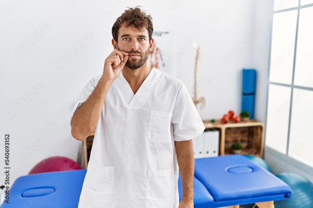 Young handsome physiotherapist man working at pain recovery clinic mouth and lips shut as zip with fingers. secret and silent, taboo talking