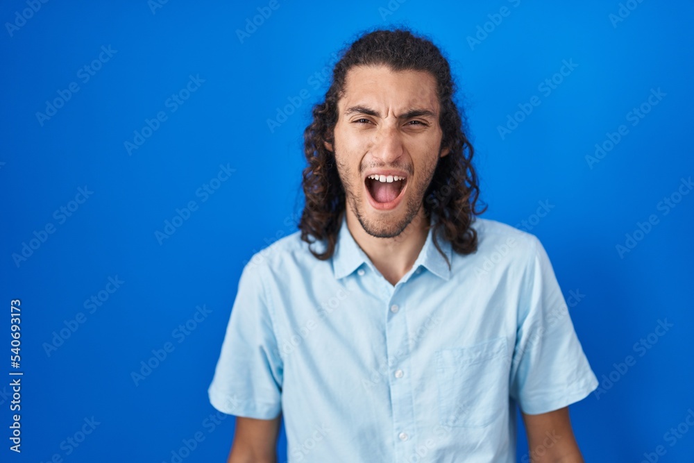 Young hispanic man standing over blue background angry and mad screaming frustrated and furious, shouting with anger. rage and aggressive concept. Young hispanic man standing over blue background angry and mad screaming frustrated and furious, shouting with anger. rage and aggressive concept.