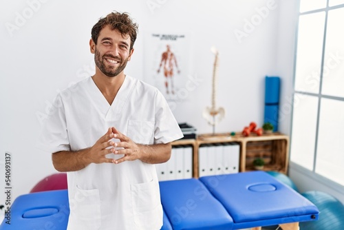 Young handsome physiotherapist man working at pain recovery clinic hands together and fingers crossed smiling relaxed and cheerful. success and optimistic
