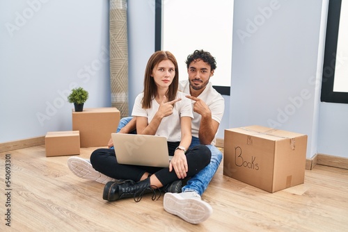 Young couple using laptop at new home pointing with hand finger to the side showing advertisement, serious and calm face