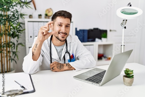 Young doctor working at the clinic using computer laptop smiling and confident gesturing with hand doing small size sign with fingers looking and the camera. measure concept.