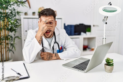Young doctor working at the clinic using computer laptop peeking in shock covering face and eyes with hand, looking through fingers with embarrassed expression.