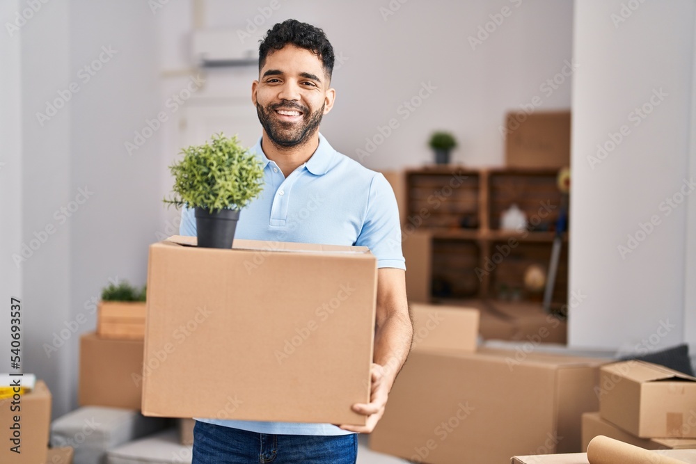 Hispanic man with beard moving to a new home holding box smiling with a happy and cool smile on face. showing teeth. Hispanic man with beard moving to a new home holding box smiling with a happy and cool smile on face. showing teeth.