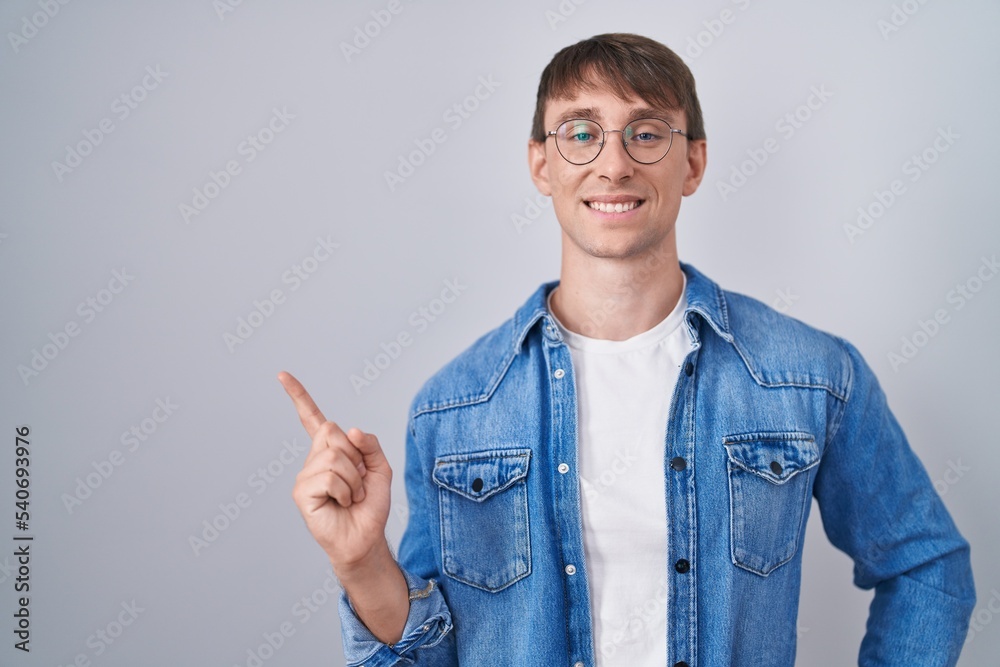 Caucasian blond man standing wearing glasses with a big smile on face, pointing with hand finger to the side looking at the camera. Caucasian blond man standing wearing glasses with a big smile on face, pointing with hand finger to the side looking at the camera.