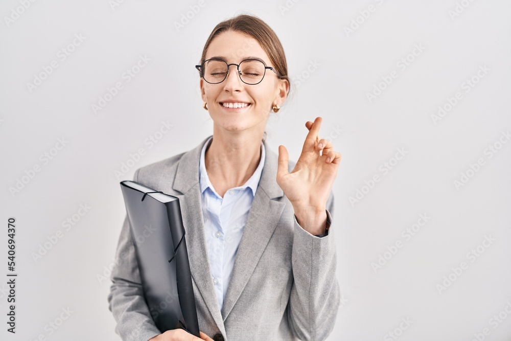 Young caucasian woman wearing business clothes and glasses gesturing finger crossed smiling with hope and eyes closed. luck and superstitious concept. Young caucasian woman wearing business clothes and glasses gesturing finger crossed smiling with hope and eyes closed. luck and superstitious concept.