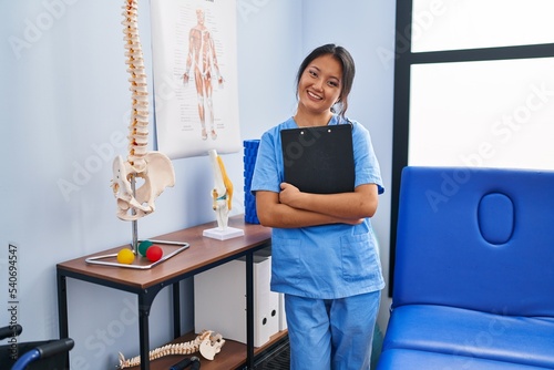 Young chinese woman wearing physiotherapist uniform holding clipboard at rehab clinic