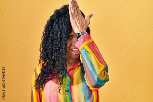 Middle age hispanic woman standing over yellow background surprised with hand on head for mistake, remember error. forgot, bad memory concept.