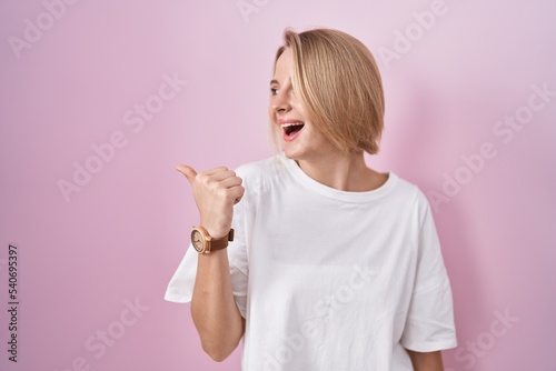 Young caucasian woman standing over pink background smiling with happy face looking and pointing to the side with thumb up. © Krakenimages.com