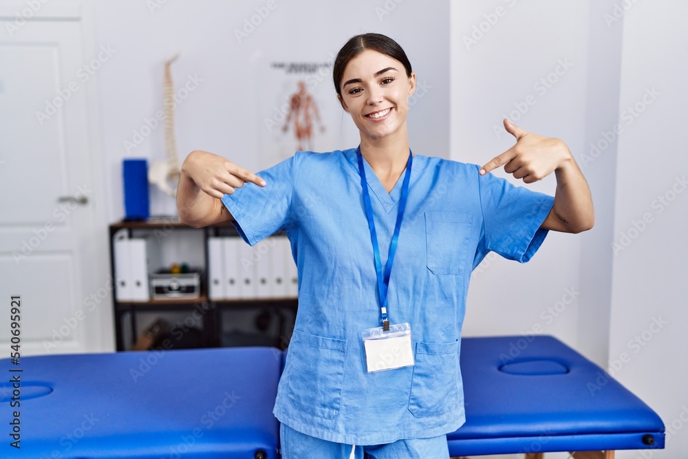 Obraz premium Young hispanic woman wearing physiotherapist uniform standing at clinic looking confident with smile on face, pointing oneself with fingers proud and happy.