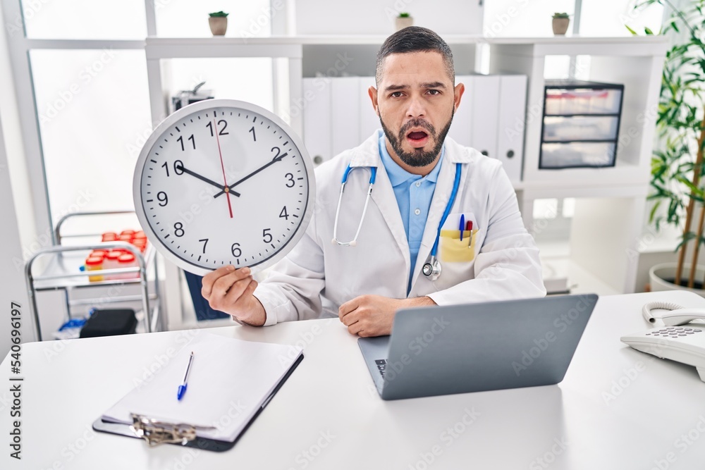 Hispanic doctor man holding clock at the clinic scared and amazed with open mouth for surprise, disbelief face Hispanic doctor man holding clock at the clinic scared and amazed with open mouth for surprise, disbelief face