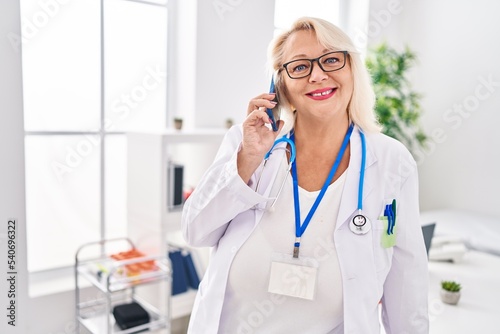Middle age blonde woman wearing doctor uniform talking on the smartphone at clinic
