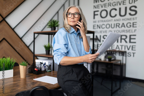 Fifty-year-old businesswoman with documents in her hands speaks on a mobile phone in a modern office. Financial Business Analytics Data Dashboard concept