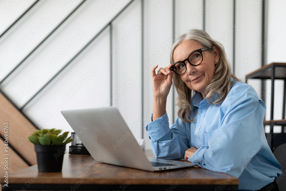 fifty-year-old businesswoman sits in front of a laptop at a table in the office and adjusts her glasses. Bank Advisor And Auditor concept fifty-year-old businesswoman sits in front of a laptop at a table in the office and adjusts her glasses. Bank Advisor And Auditor concept