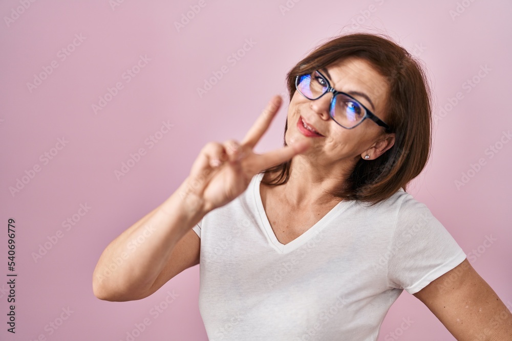 Middle age hispanic woman standing over pink background smiling looking to the camera showing fingers doing victory sign. number two.