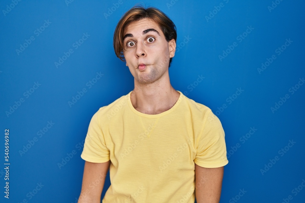 Young man standing over blue background making fish face with lips, crazy and comical gesture. funny expression.