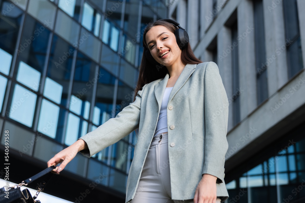 Happy young woman in smart suit wearing headphones looking at camera smiling holding bag in hand relaxing after hard day standing near buisness centre posing for camera.