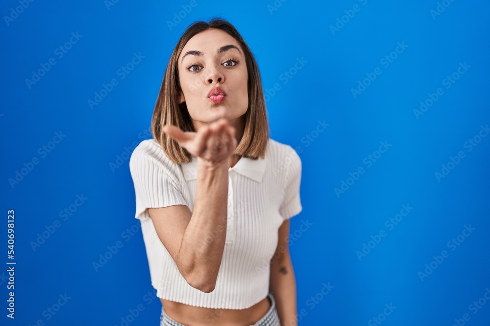 Hispanic woman standing over blue background looking at the camera blowing a kiss with hand on air being lovely and sexy. love expression. Hispanic woman standing over blue background looking at the camera blowing a kiss with hand on air being lovely and sexy. love expression.