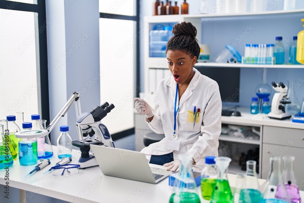 Young african american with braids working at scientist laboratory with laptop scared and amazed with open mouth for surprise, disbelief face Young african american with braids working at scientist laboratory with laptop scared and amazed with open mouth for surprise, disbelief face