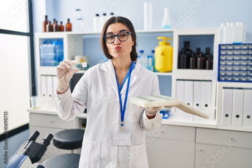 Young hispanic woman working at scientist laboratory with blood samples looking at the camera blowing a kiss being lovely and sexy. love expression.