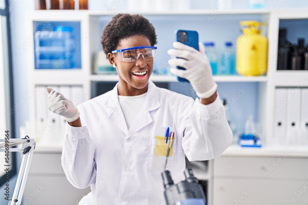 African american woman working at scientist laboratory doing video call with smartphone screaming proud, celebrating victory and success very excited with raised arm African american woman working at scientist laboratory doing video call with smartphone screaming proud, celebrating victory and success very excited with raised arm