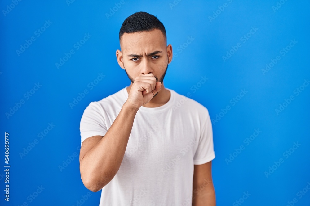 Young hispanic man standing over blue background feeling unwell and coughing as symptom for cold or bronchitis. health care concept. Young hispanic man standing over blue background feeling unwell and coughing as symptom for cold or bronchitis. health care concept.