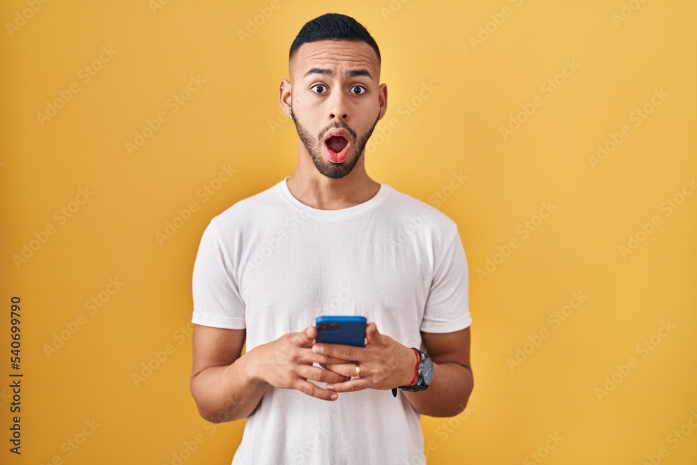 Young hispanic man using smartphone typing message afraid and shocked with surprise and amazed expression, fear and excited face.