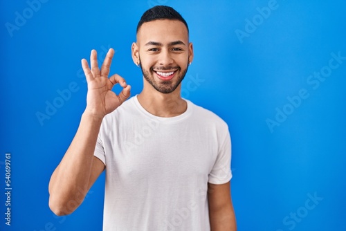 Young hispanic man standing over blue background smiling positive doing ok sign with hand and fingers. successful expression.