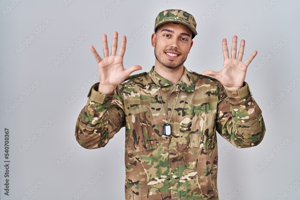 Young hispanic man wearing camouflage army uniform showing and pointing up with fingers number ten while smiling confident and happy. Young hispanic man wearing camouflage army uniform showing and pointing up with fingers number ten while smiling confident and happy.