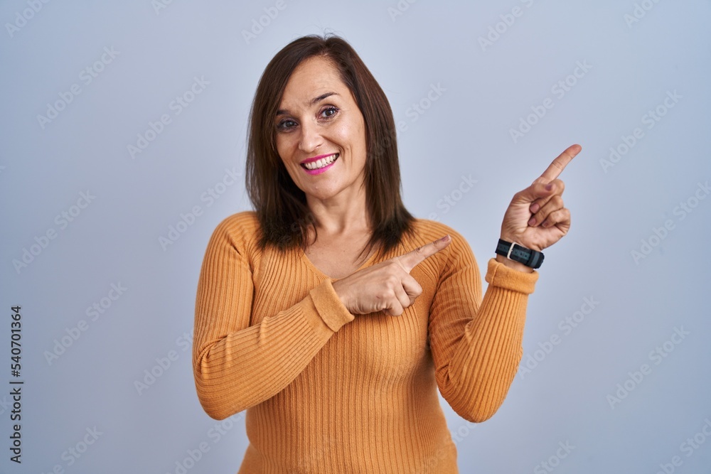 Middle age brunette woman standing wearing orange sweater smiling and looking at the camera pointing with two hands and fingers to the side. Middle age brunette woman standing wearing orange sweater smiling and looking at the camera pointing with two hands and fingers to the side.